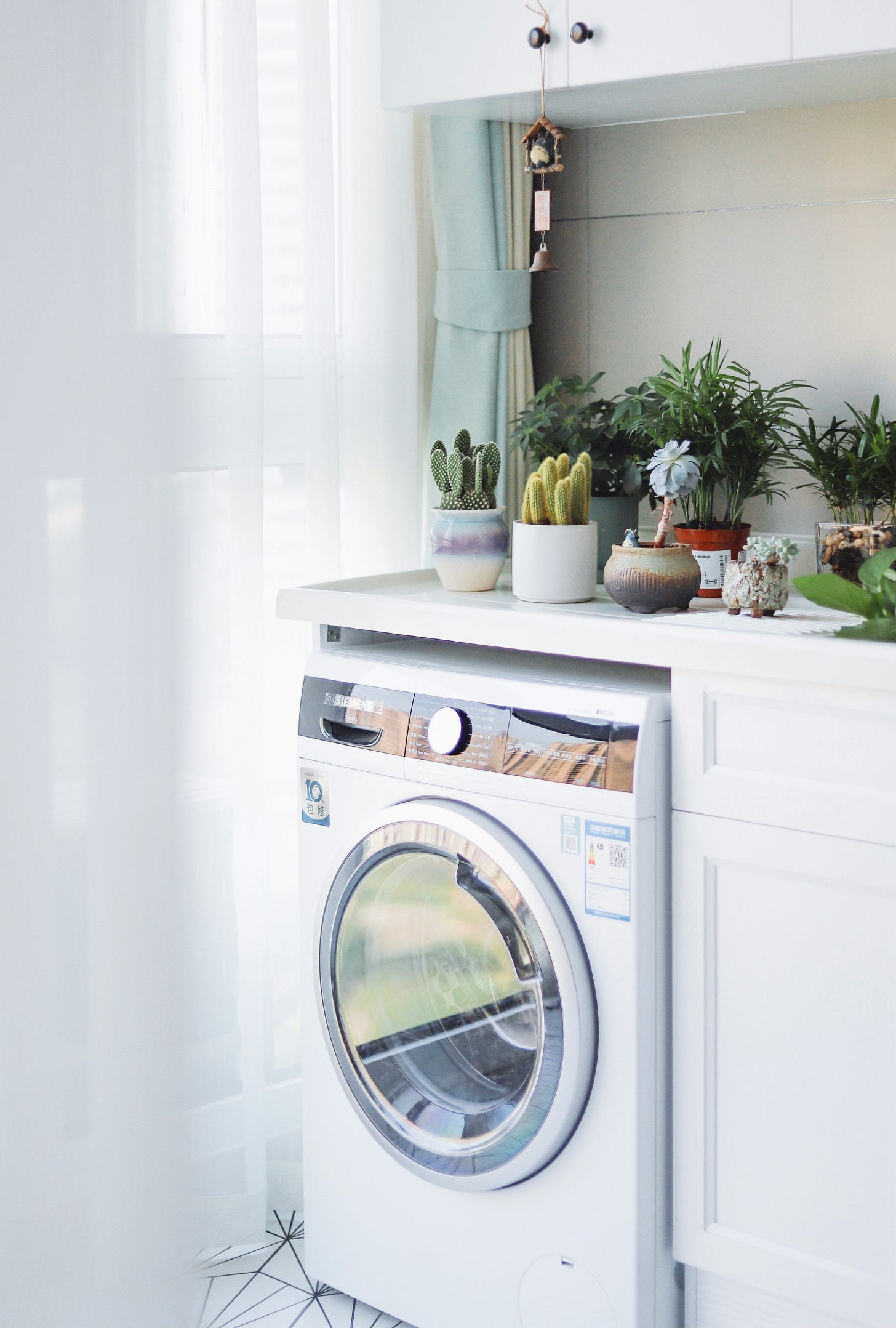 A washing machine/dryer unit sitting in a house below the countertop. Several small pots of plants sit on top of the counter. Next to the machine is a white curtain. A washing machine/dryer unit sitting in a house below the countertop. Several small pots of plants sit on top of the counter. Next to the machine is a white curtain.