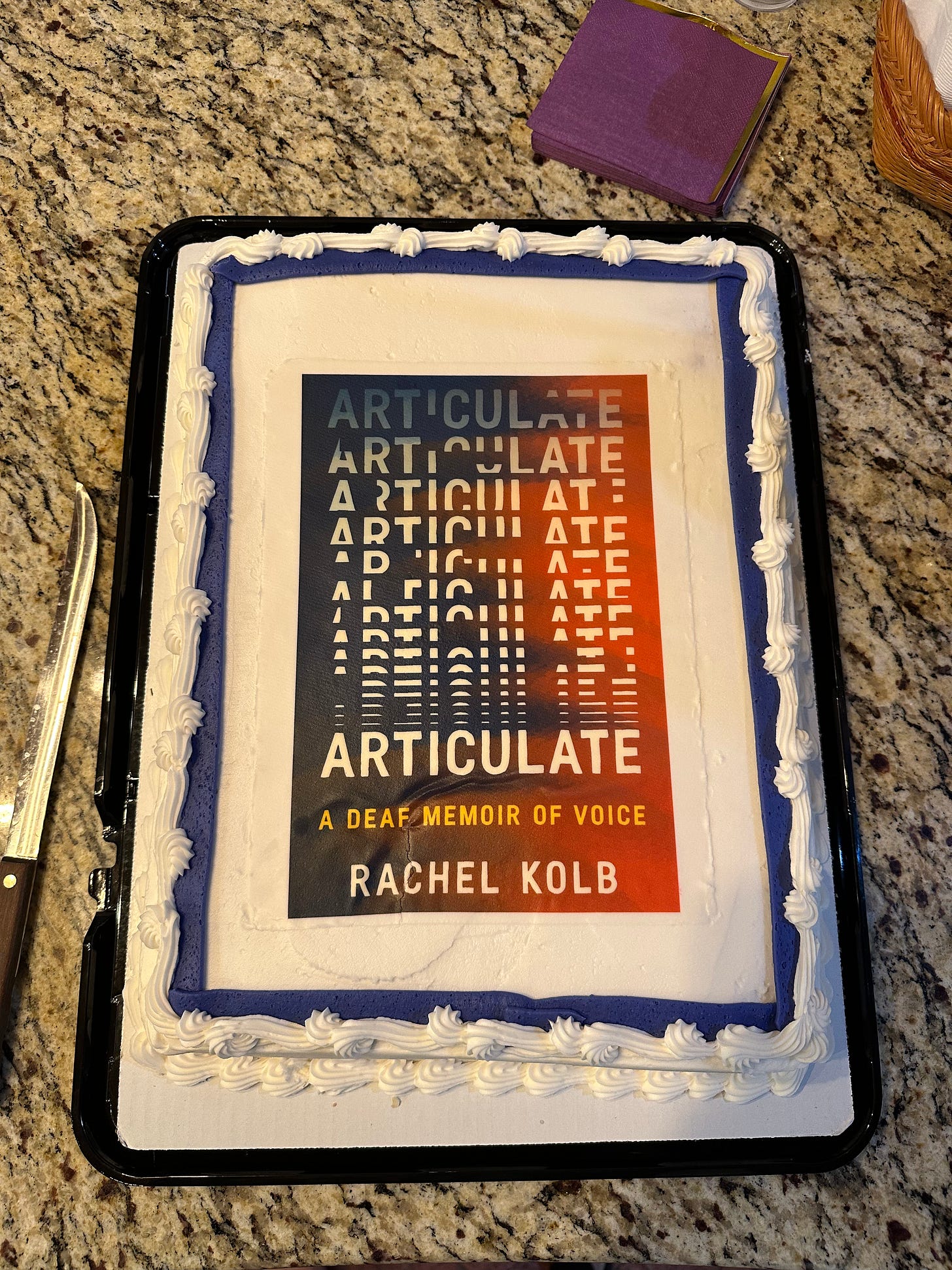 Photo of a white sheet cake with purple border, laid against a gray marble countertop with napkins and knives. On the top of the cake is an image of my book ARTICULATE, the letters of the title gradually assembling across the page as purple-and-pink ripples swirl in the background.