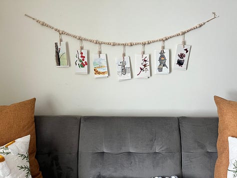 A series of mixed-media cards hang on a beaded string and in a frame, a painting of a shipwreck ocean scene with a wooden frame, an abstract view through my dining room window of the mountain carved into a wooden cutting board, and a patchwork quilt laying atop a couch.