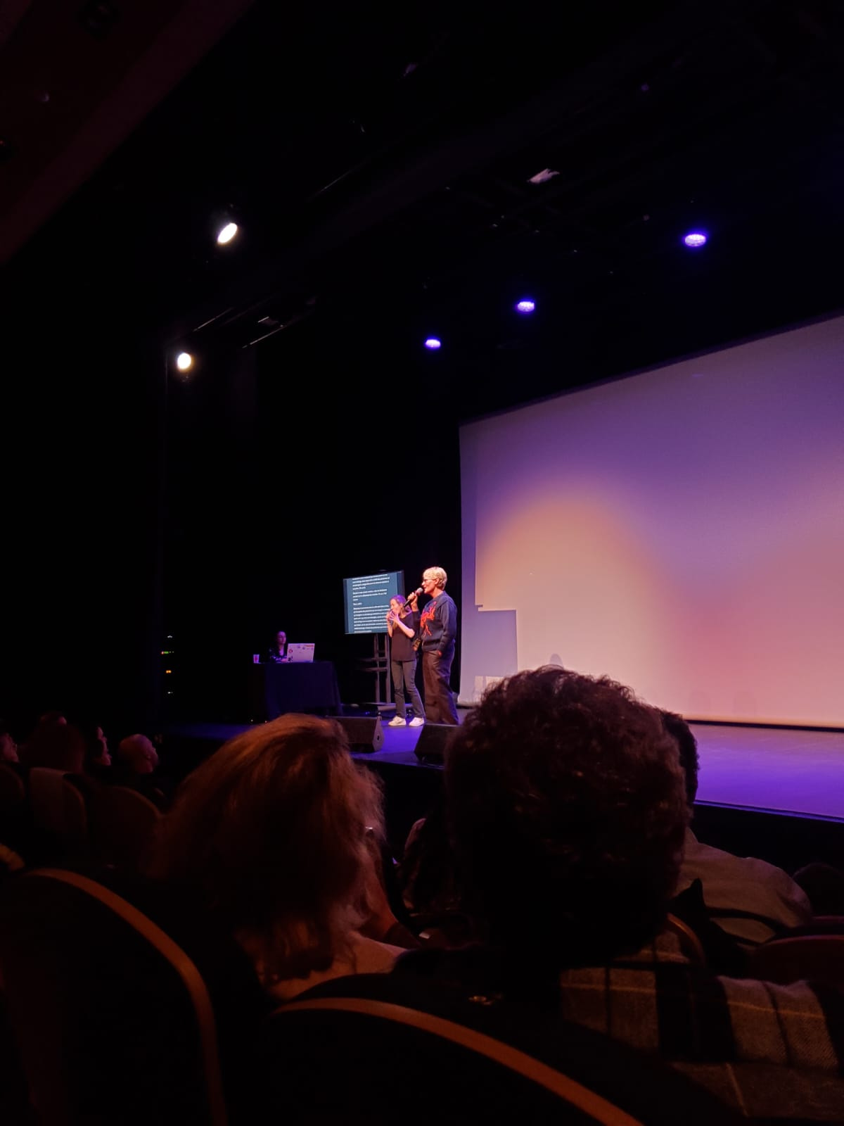 Sur une scène illuminée en violet, Judith Godrèche parle dans un micro. A côté d’elle, Marina, tout en noir, interprète en langue des signes et à côté d’elle, Céline tape sur son ordinateur. Ce qu’elle écrit est retranscrit sur un écran placé à côté de Marina.