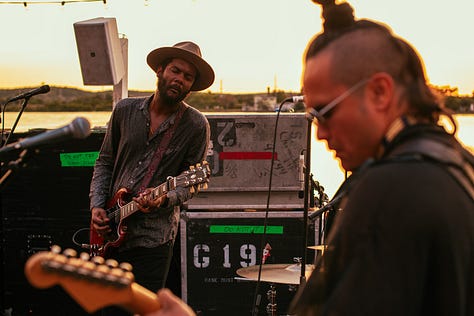 A gallery of three photos of Gary Clark Jr. and his band performing on an outdoor stage. The left photo shows him in silhouette against a bright orange sun, the middle photo shows him playing guitar, and the right photo shows him playing next to his bassist.