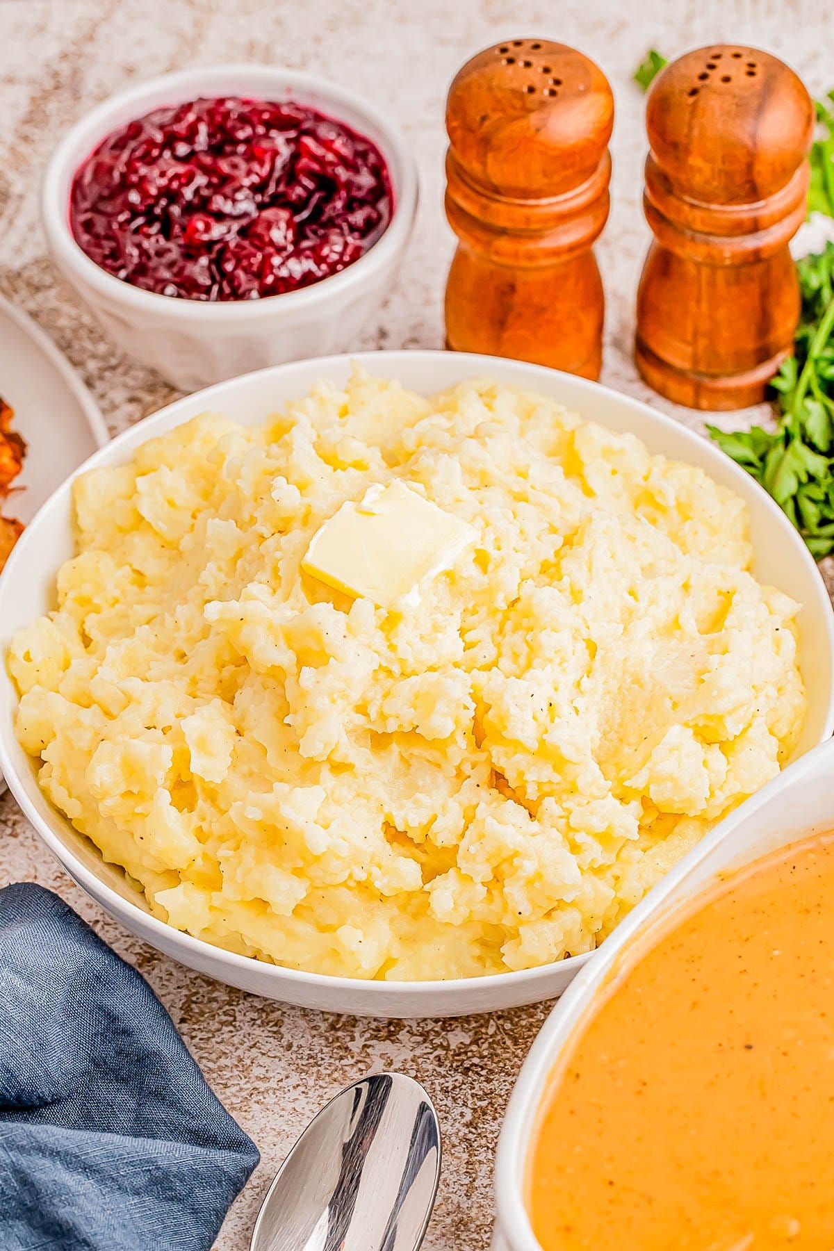 A bowl of mashed potatoes with butter on top, surrounded by bowls of cranberry sauce and gravy, with salt and pepper shakers in the background. A bowl of mashed potatoes with butter on top, surrounded by bowls of cranberry sauce and gravy, with salt and pepper shakers in the background.