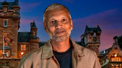 A man in a dark t-shirt and light brown shirt smiles to camera, set against the backdrop of a castle by night A man in a dark t-shirt and light brown shirt smiles to camera, set against the backdrop of a castle by night