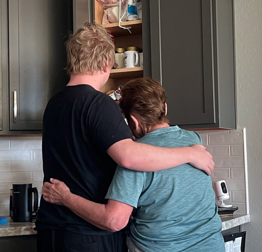 two people facing a kitchen cabinet, arms around each other