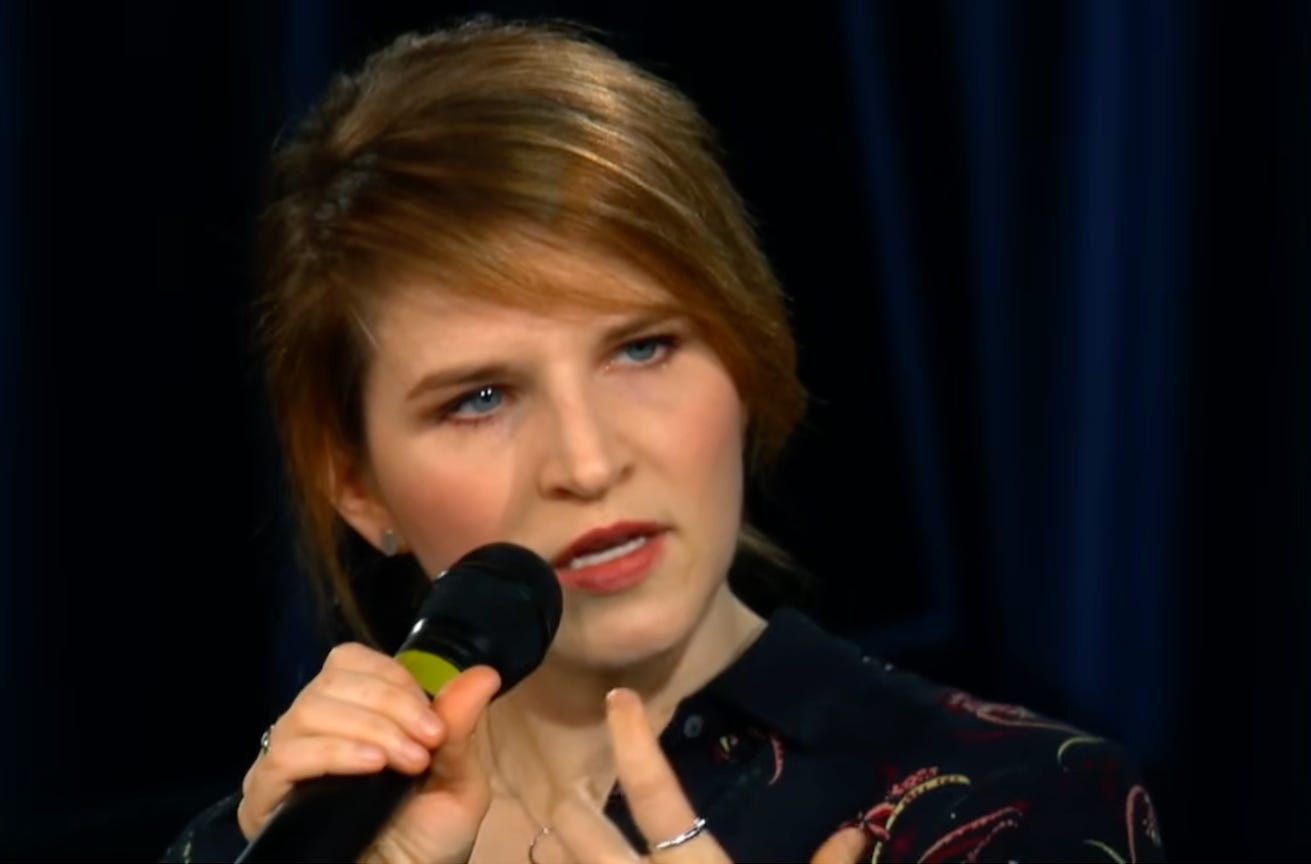 Headshot of Tara Westover in an interview, holding a microphone, gesturing with her free hand.