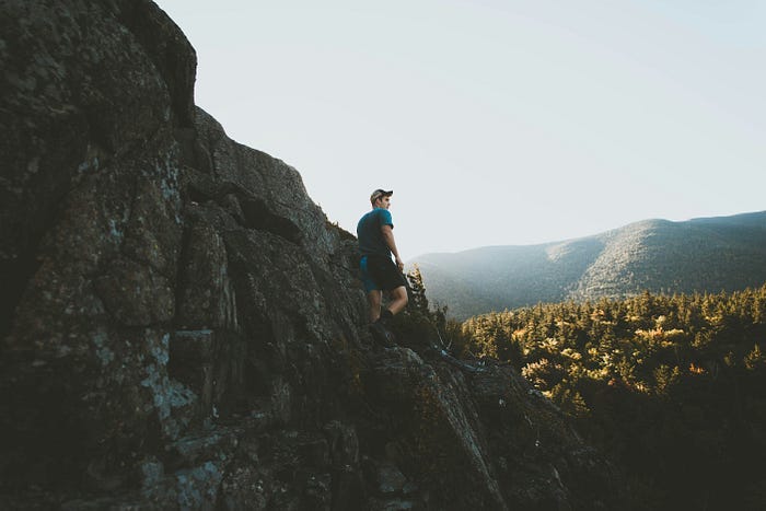A man standing on cliff face with t-shirt and a hat, looking over a wide forest and mountain range in the background