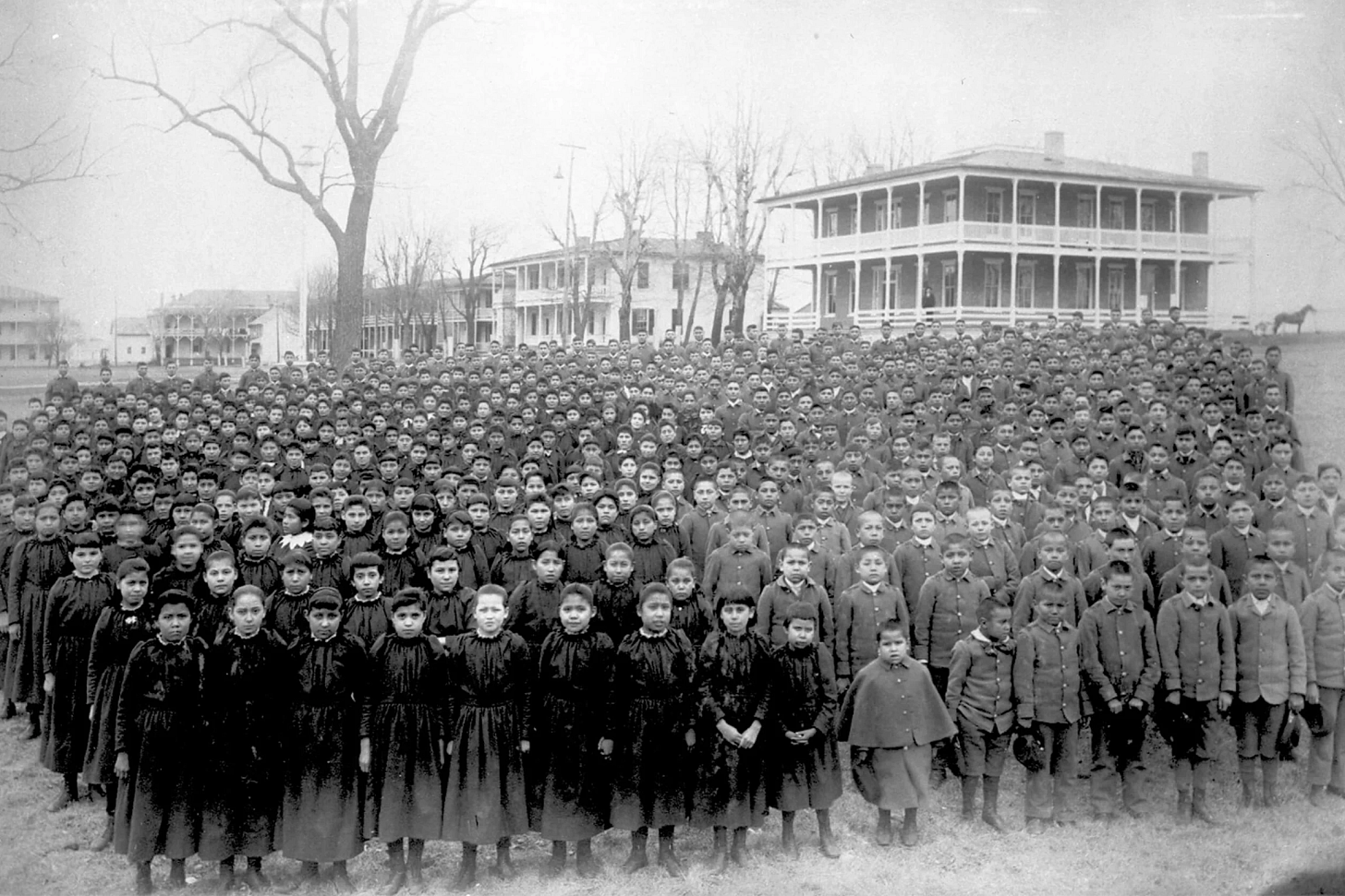 In a historical photo, a group of Native American students dressed in school uniforms standing in an open lawn
