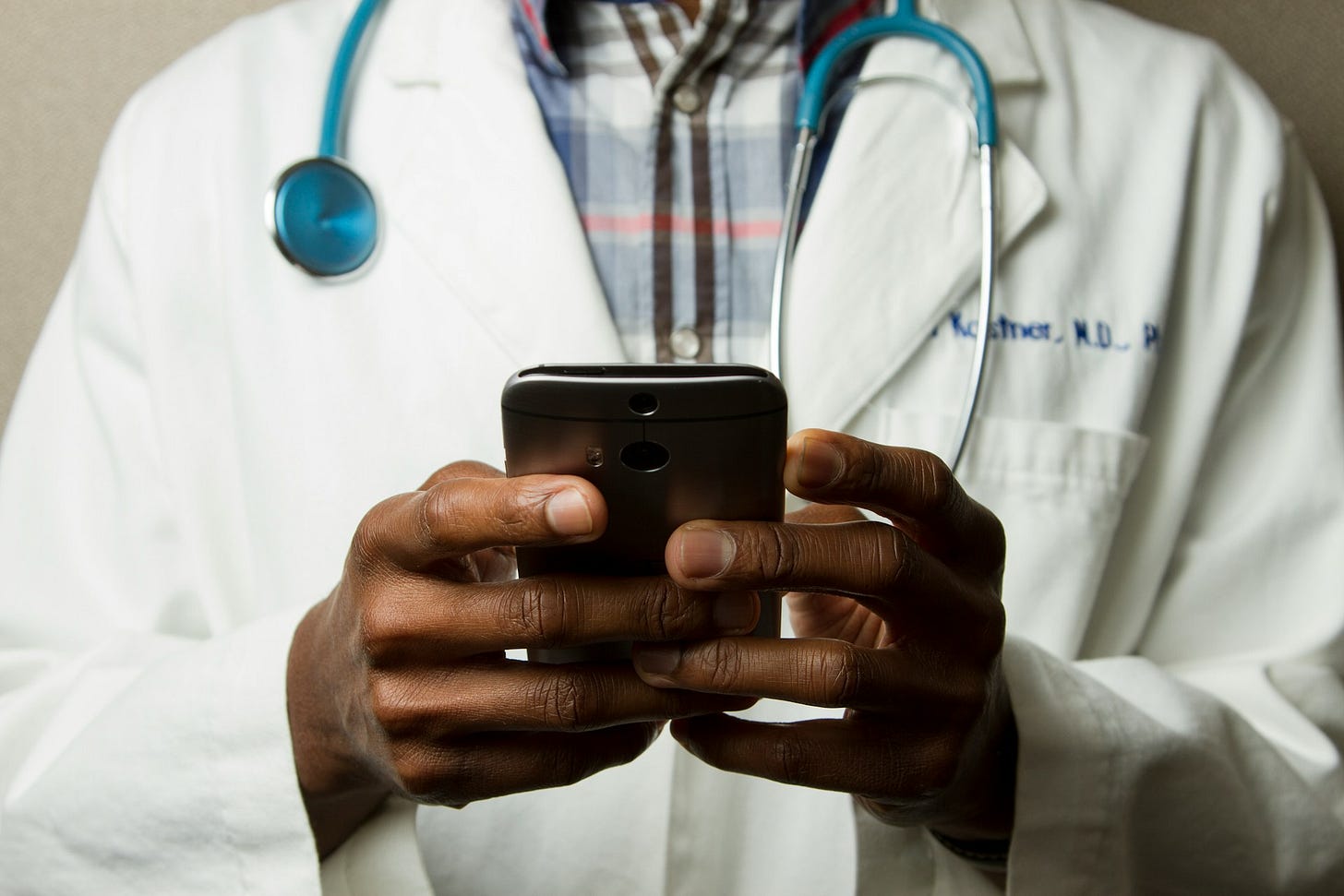 A doctor in a white coat and stethoscope holds a smartphone, symbolizing healthcare professionals using social media to connect with patients. Photo by National Cancer Institute on Unsplash