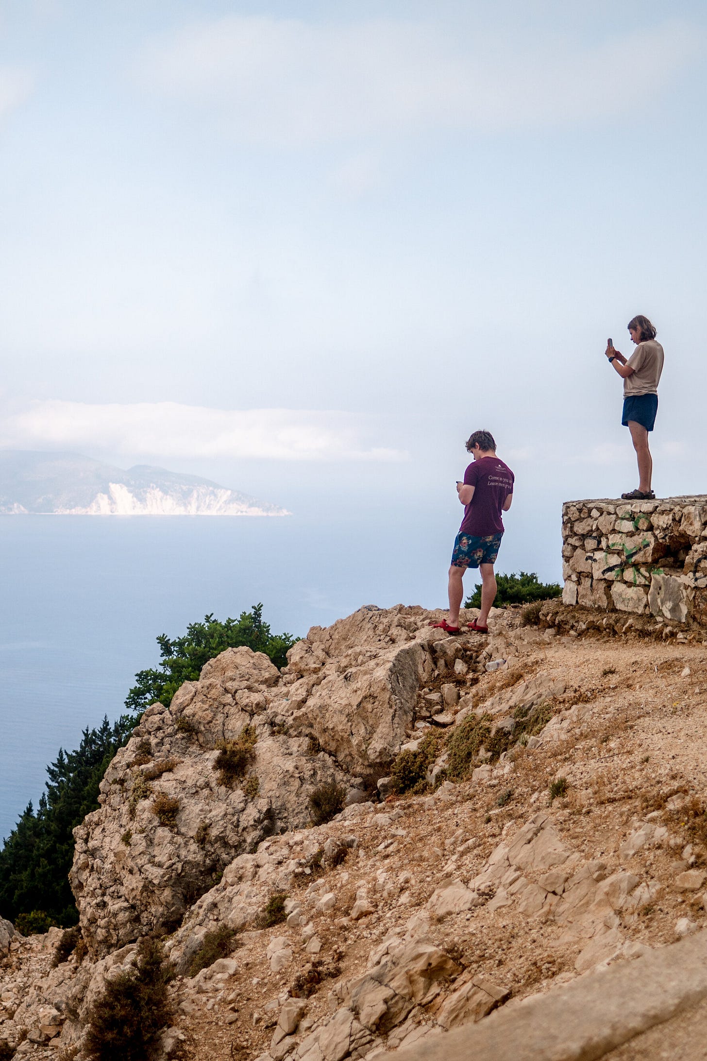 Two people take photos using their phones on a rocky cliff overlooking a vast body of water.