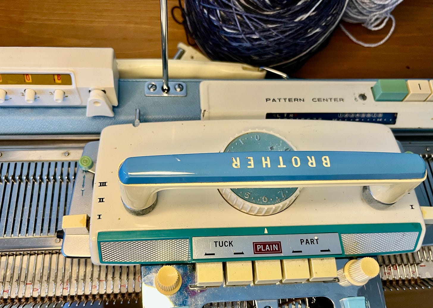 Close-up of a Brother KH585 knitting machine carriage from the mid-1960s. The blue and cream carriage shows TUCK, PLAIN, and PART stitch selector buttons. A transfer tool rests on the needle bed. The Pattern Center housing is visible at the right.