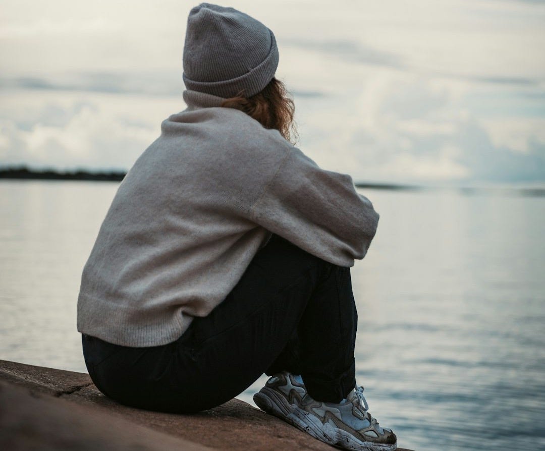 person in gray hoodie and black pants sitting on brown wooden dock during daytime