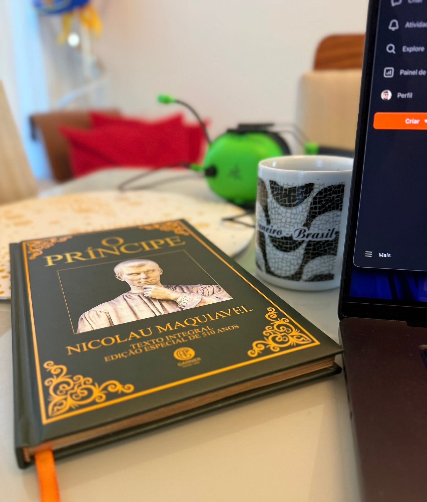 Livro O Príncipe, edição ouro da editora Garnier, próximo a um notebook e um café, sob uma mesa de jantar. Livro O Príncipe, edição ouro da editora Garnier, próximo a um notebook e um café, sob uma mesa de jantar.