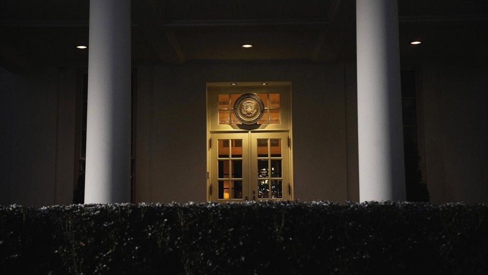 A color photograph of a White House door at night, seen behind a hedge and between two columns.