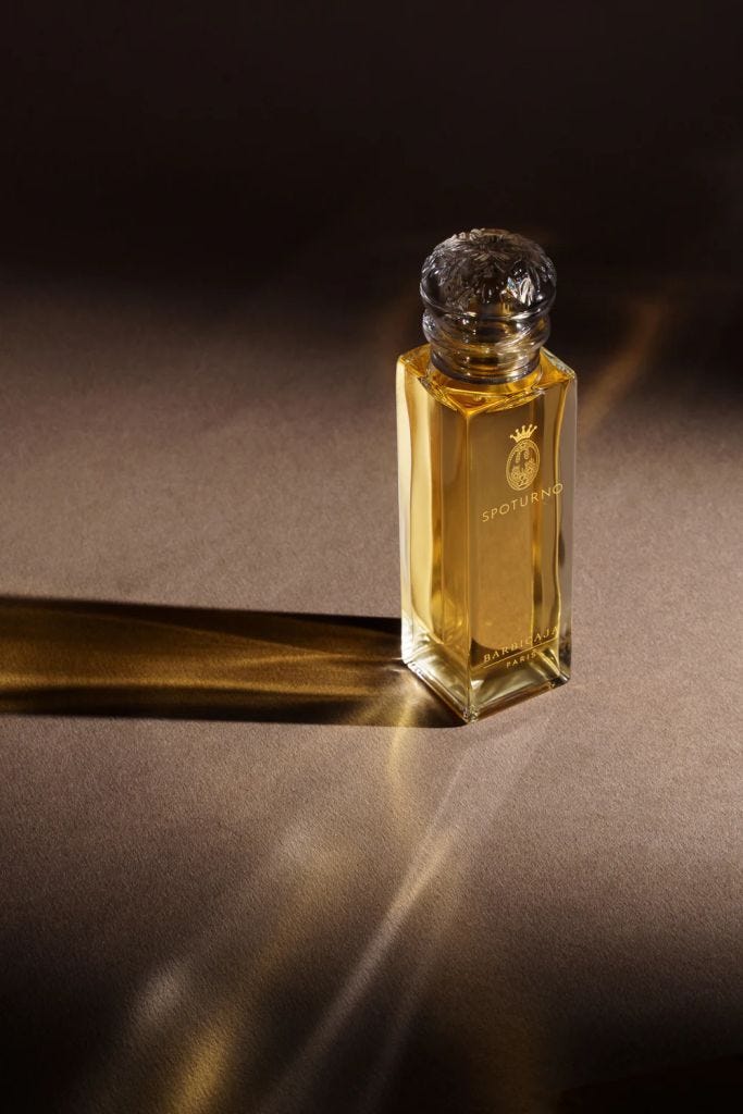 A glass perfume bottle with a ornate cap, filled with a golden liquid, casting soft shadows on a textured surface.