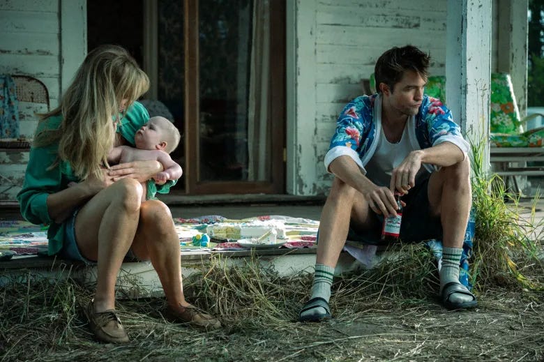 Lawrence and Pattison sitting on stoop of their run down house. Lawrence has her face covered by her hair as she looks down at the baby; Pattison has legs spread holding a beer between them. They are both in sloppy states of near undress