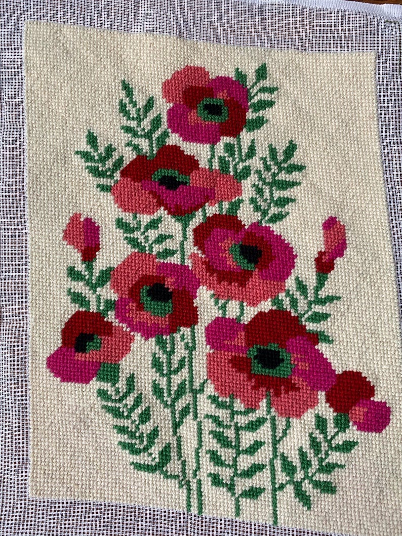 May include: Needlepoint art featuring a floral design. The artwork showcases a bouquet of flowers in shades of red and pink, with green leaves and stems. The flowers have dark centers, and the background is a cream color. The piece is framed by a white mesh.