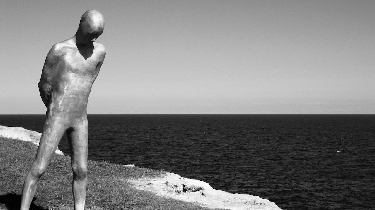 Black and white photo of a minimalist human-like sculpture standing on a grassy cliffside overlooking the ocean. The figure is slightly hunched forward with arms behind its back, facing downwards toward the ground, evoking a contemplative or solemn mood. The sea stretches out to the horizon under a clear sky.