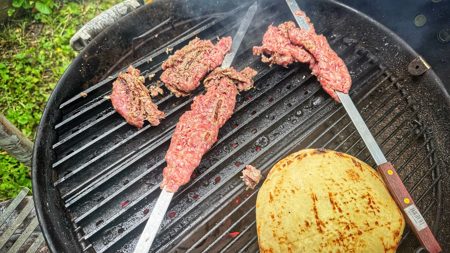 The koobideh skewers falling apart on the grill . Two skewers still have some meat clinging to them but one has lost all of its meat and is out of the photo. On the cool side of the grill, some naan is warming.