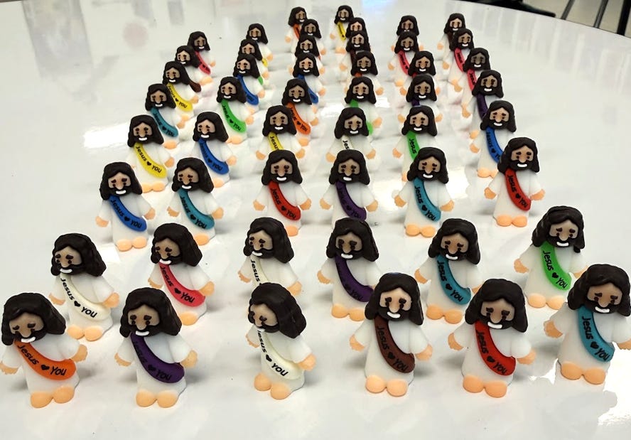 Dozens of small plastic Jesus figurines stand arranged in neat rows across a white tabletop. Each figure wears a white robe and a brightly colored sash reading “Jesus Loves You,” with arms extended in a welcoming pose. The collection resembles a tiny congregation and represents the bulk purchase that eventually led to fifty-four miniature Jesuses taking up residence on the author's classroom whiteboard. Dozens of small plastic Jesus figurines stand arranged in neat rows across a white tabletop. Each figure wears a white robe and a brightly colored sash reading “Jesus Loves You,” with arms extended in a welcoming pose. The collection resembles a tiny congregation and represents the bulk purchase that eventually led to fifty-four miniature Jesuses taking up residence on the author's classroom whiteboard.