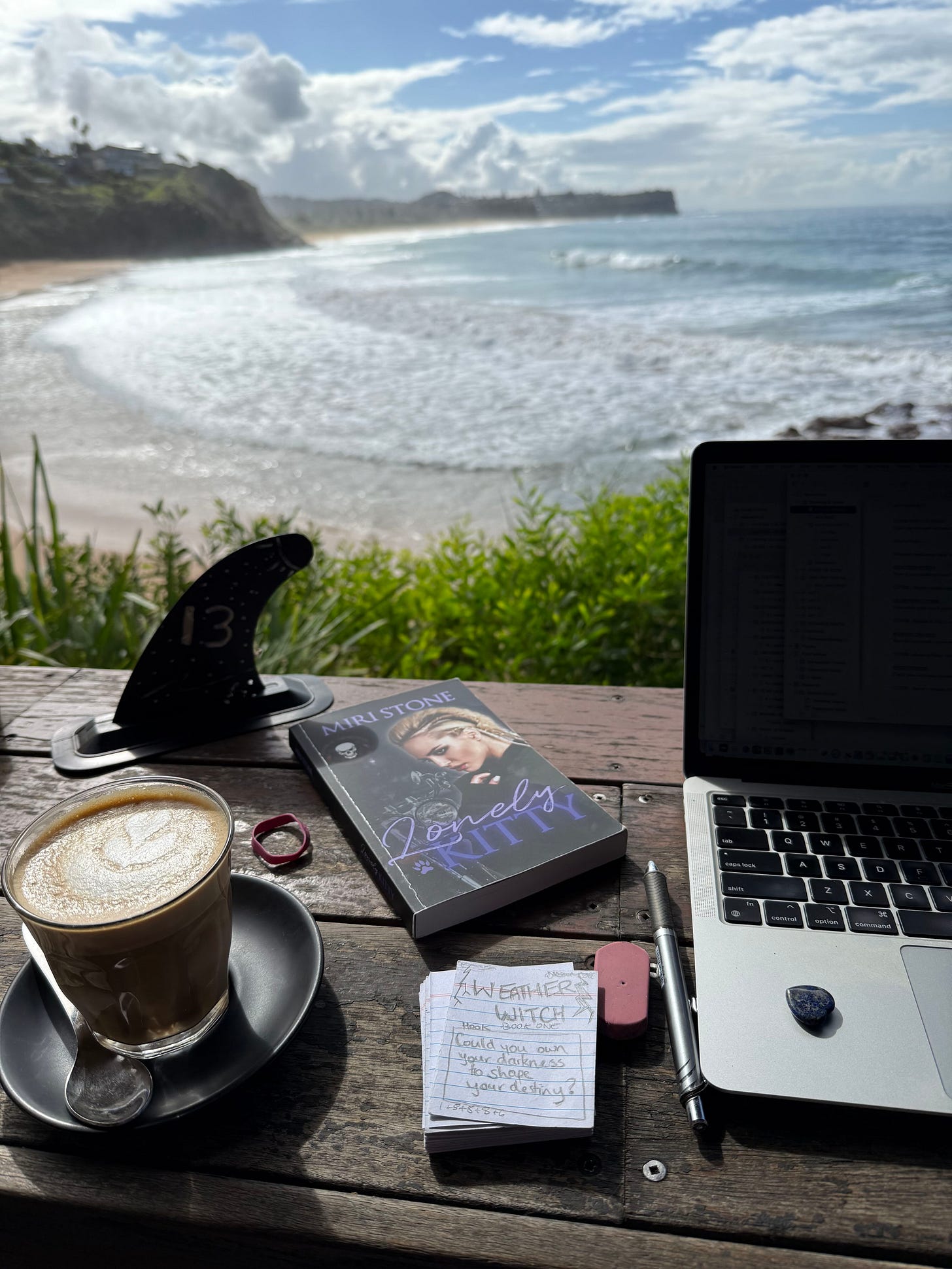 Photo taken from Miri's point of view of working at a favorite cafe with her laptop, a paperback copy of Lonely Kitty, and her beat cards for Weather Witch. Stretched out in the background is a stunning view of Warriewood surf beach.