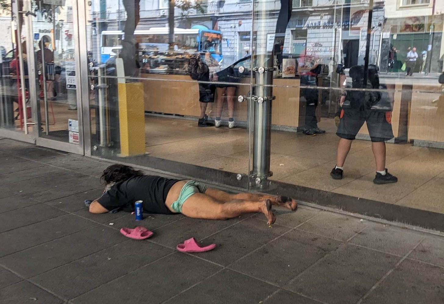a man lying on a sidewalk in underwear and a T-shirt with a can of beer next to him.