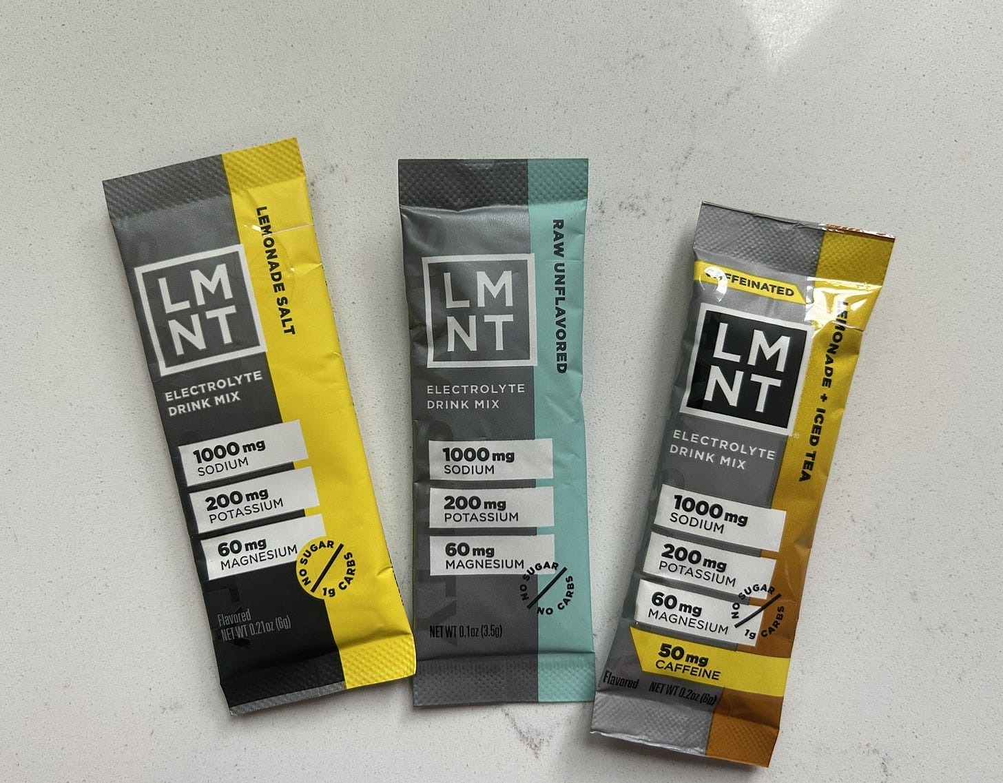 Three LMNT electrolyte stick packets on a counter: Lemonade Salt, Raw Unflavored, and caffeinated Lemonade and Iced Tea. Three LMNT electrolyte stick packets on a counter: Lemonade Salt, Raw Unflavored, and caffeinated Lemonade and Iced Tea.