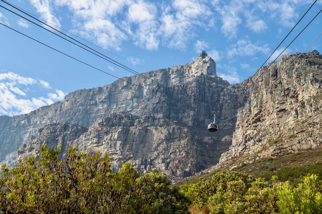 A cable car ascends toward the sandstone cliffs of Table Mountain in Cape Town, with support cables crossing a partly cloudy sky and fynbos shrubs in the foreground. A cable car ascends toward the sandstone cliffs of Table Mountain in Cape Town, with support cables crossing a partly cloudy sky and fynbos shrubs in the foreground.
