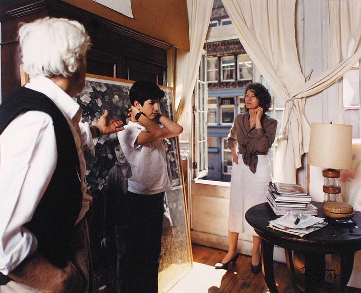 1: Photograph by Tina Barney depicting a bed covered in a white embroidered bedspread adorned with pink flowers. A young girl in a white dress with puffed sleeves and her hair adorned with ribbons sits on the bed. Next to her is a man in a blue shirt, leaning on the bed, appearing to engage in conversation with her. In the background, another girl in a pastel-colored dress stands in a brightly lit closet filled with clothes hanging on a rod. 2: Photograph by Tina Barney depicting two women in a room with cream colored walls. On the left, one woman with wavy brown hair is seated, turned toward the camera, resting her chin on her hand. To the right, facing a mirror, a young woman with short, bleach blonde hair applies bright red lipstick. She is also dressed in black and stands in a profile position, reflecting in the mirror. 3: Photograph by Tina Barney, depicting three people engaged in conversation in a sunlit room. On the left, an older man with white hair wearing a black vest over a white shirt gestures towards the center. In the middle, a young person stands with their hands holding the back of their neck. To the right, a woman in a light brown blouse and white skirt holds her chin thoughtfully. 4: Photograph by Tina Barney featuring two women in a pastel pink bathroom, both wearing pink robes. On the left, one woman with short dark hair and glasses gazes toward the camera, her hand on her hip. She is standing in front of a mirror, in which her reflection is visible. To the right, a young woman with long dark hair peeks from behind floral curtains covering a window.