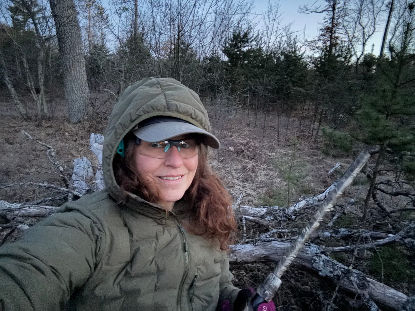 A woman wearing all olive drab is seated on a down tree in the woods in the early morning, holding a shotgun and wearing safety glasses under her baseball cap and hood. She is smiling tentataively at the camera even though she is half-frozen and hasn't heard a turkey.