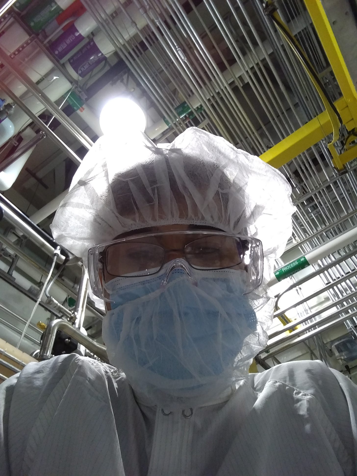 A person wearing a cleanroom suit, safety glasses, and a mask beneath manufacturing equipment