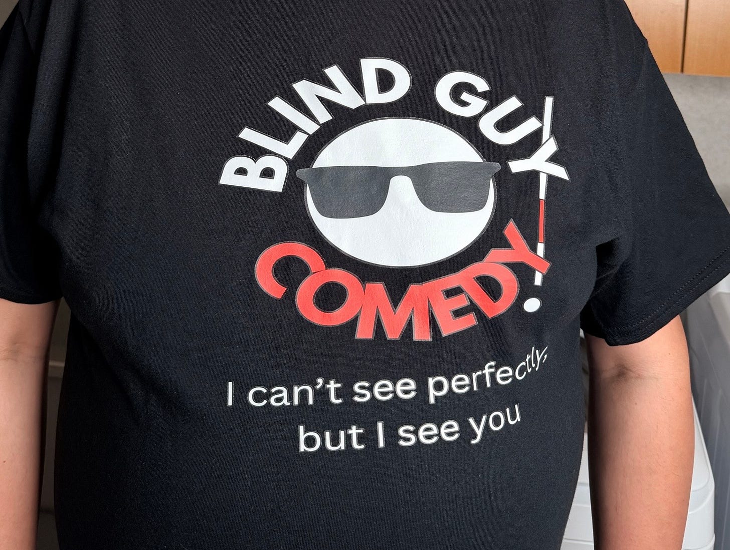 A close-up photo of a person wearing a black Blind Guy Comedy T-shirt. The design features a white circular face wearing black sunglasses, with the words “BLIND GUY” in white above and “COMEDY!” in red below. A white cane forms the exclamation mark beside the logo. Beneath the graphic, the slogan reads, “I can’t see perfectly, but I see you.” The image focuses on the shirt design, with only the wearer’s torso and arms visible.