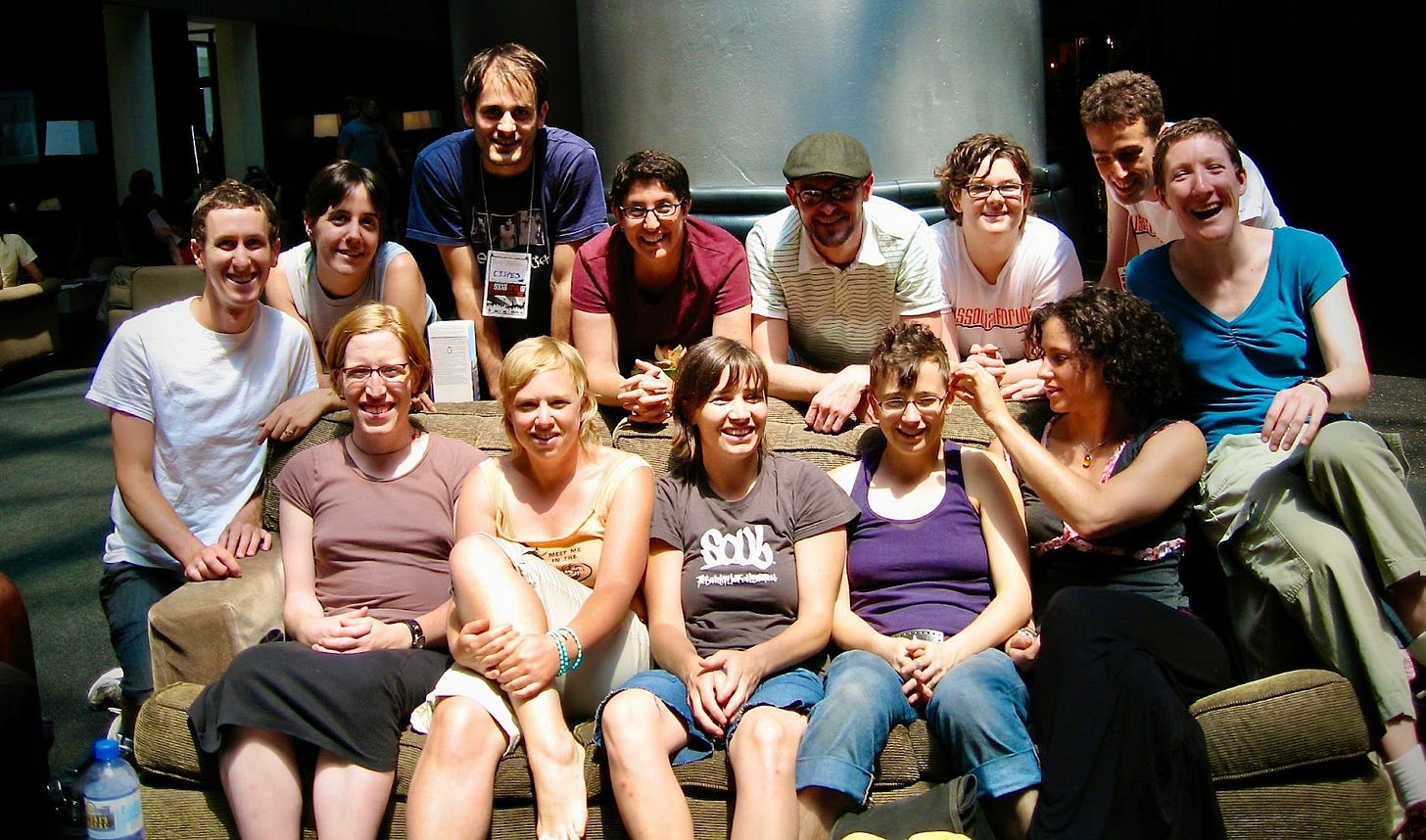 A group photo of a bunch of white, casually dressed, young adults sitting and standing and crouching on and around a couch. Everyone is smiling for the camera.