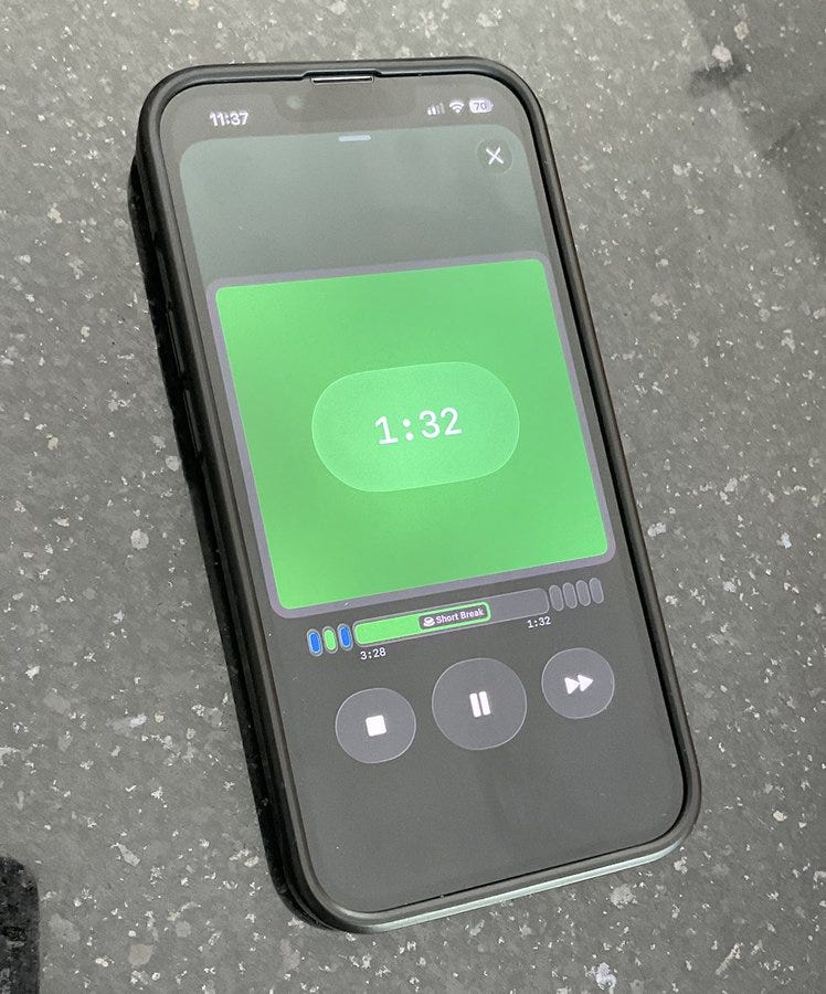A black smartphone displaying a green Pomodoro timer app interface. The screen shows a green background with a timer reading 1:32, play/pause controls, and a progress bar below. The phone is placed on a dark, speckled surface.