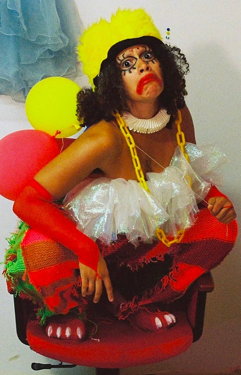 Chimera the Clown has light brown skin and wears hand knotted red and green woven pants wtih red clawed, croc-style shoes poking undernearth. The wear a chiffon, iradescent clown collar as a top, with a small Elizabeathan collar and plastic yellow chain as a necklace. They wear a fuzzy, tall yellow hat and their face is painted with an exaggerated red clown frown, and much wider than realistic 60's inspired make up with a cartoonish outline of the eye with big eyeslashes. Their hair is big, dark, and curly. They hold deflating red and yellow balloons with a light blue tutu and a white wall behind them. In the center protrait, they have edited the photo to make themself look like a clown-fairy with blue skin, a long blue fish-like tail and blue butterfly wings. They are standing in a tea cup on a table of many colorful tea cups. The outter two images are them crouching on a red desk chair. One with their arms down. The other with their arms raised. The pants and image are by GAUDMOTHER. The styling, make up, and edit is Malachi Lily. 