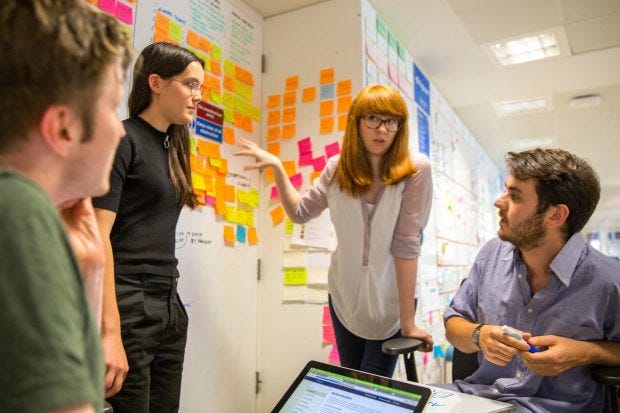 Two young female and two male designers discuss ideas in front of a wall with colour stickie notes; one holds a laptop computer, another one has a sketchpad in front of him Two young female and two male designers discuss ideas in front of a wall with colour stickie notes; one holds a laptop computer, another one has a sketchpad in front of him