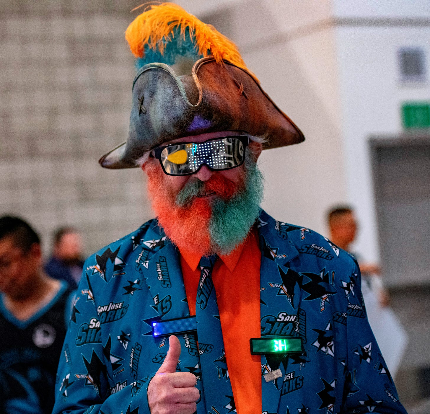 Man wearing a Sharks jacket, with a tie dyed beard, and a feathered pirate hat