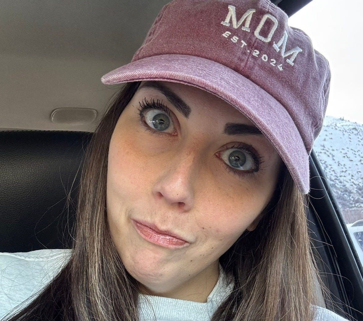 Katlyn Slone, wearing a mauve baseball cap that reads “MOM EST. 2024,” gives a playful, wide-eyed expression while sitting in a car. Her raised eyebrows and slight smirk hint at disbelief and pride. Katlyn Slone, wearing a mauve baseball cap that reads “MOM EST. 2024,” gives a playful, wide-eyed expression while sitting in a car. Her raised eyebrows and slight smirk hint at disbelief and pride.