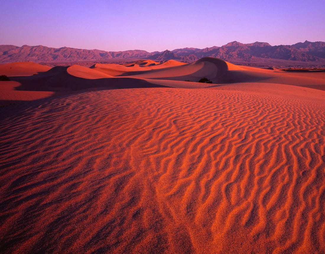 Wind-sculpted sand dunes glowing red and orange at sunset in Death Valley, with rippled textures in the foreground and distant mountains under a purple sky Wind-sculpted sand dunes glowing red and orange at sunset in Death Valley, with rippled textures in the foreground and distant mountains under a purple sky