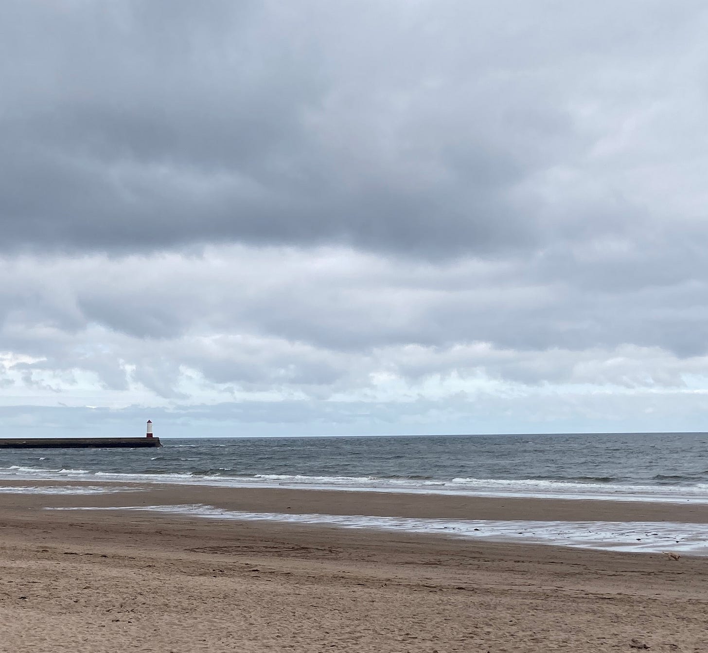 Grey atmosphere of North Sea with large expanse of cloud above, and a lighthouse in the middle distance with an expanse of beach in the foreground