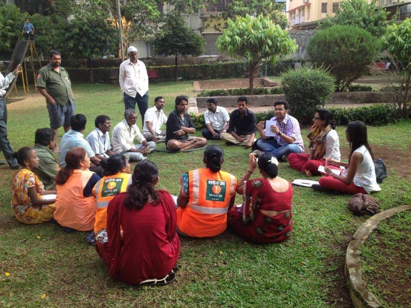 A group of 18 Indian people sit in a circle in a part, while several men stand in the background.