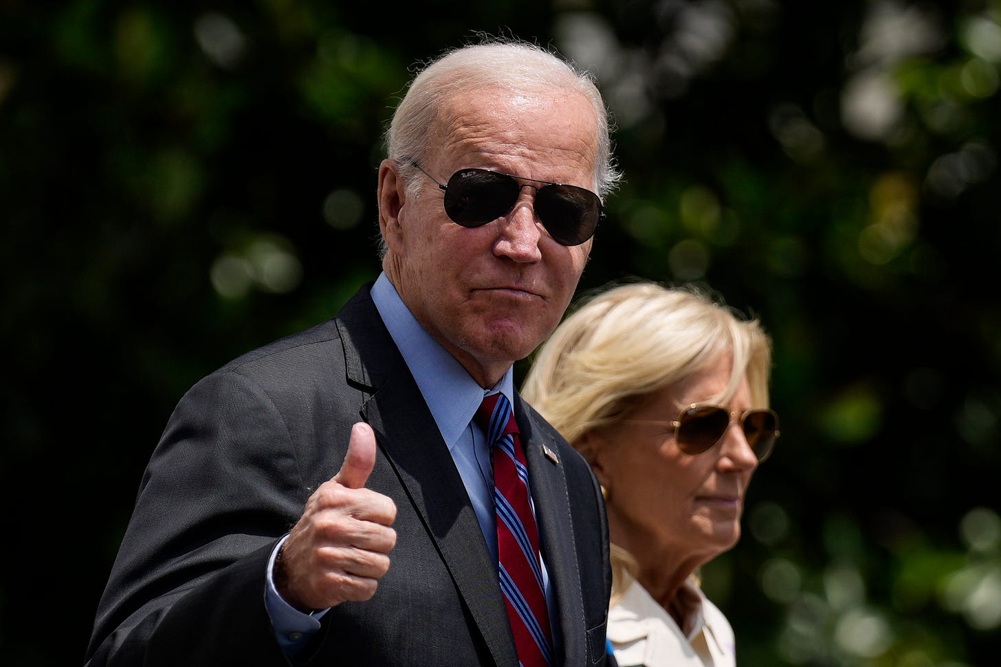 An older white man with white hair wearing a dark suit, sunglasses and a blue and red tie gives a thumbs up, a white woman with blonde hair and sunglasses is walking behind him An older white man with white hair wearing a dark suit, sunglasses and a blue and red tie gives a thumbs up, a white woman with blonde hair and sunglasses is walking behind him