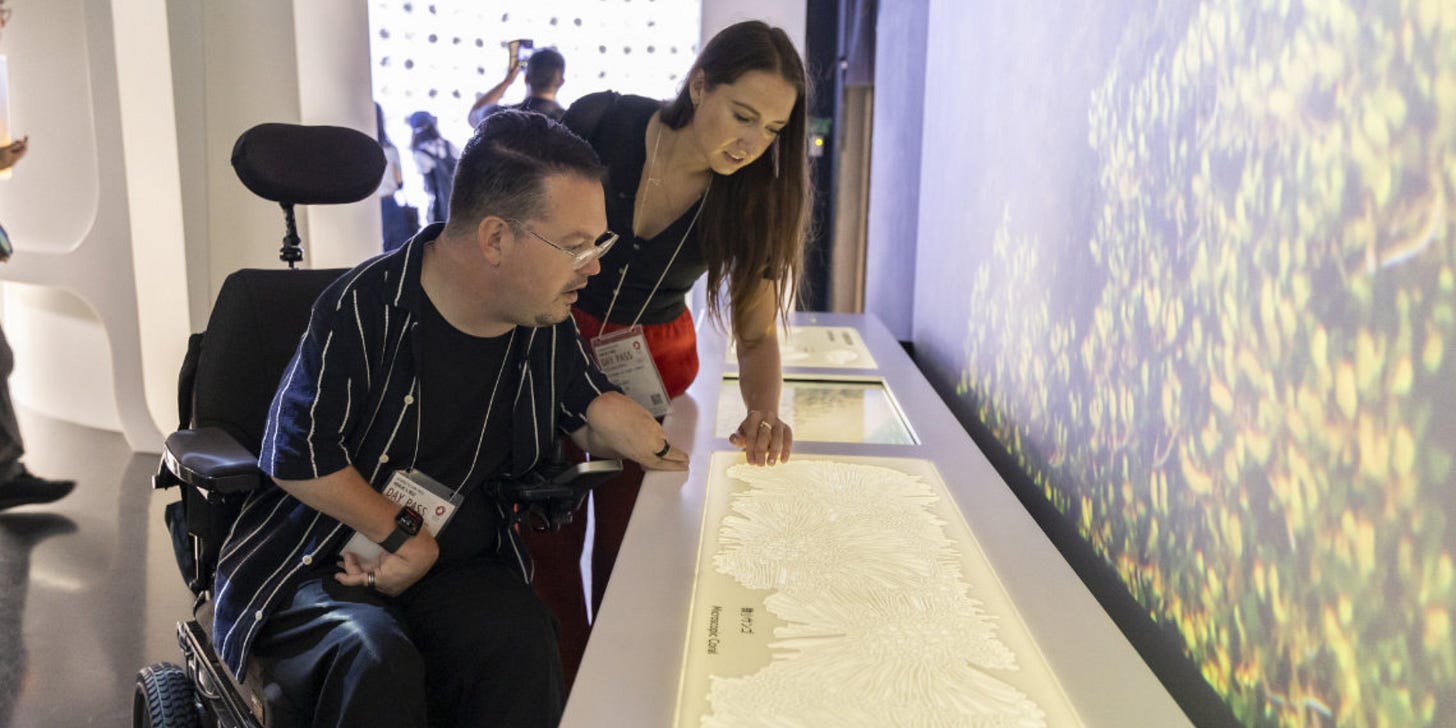 Josh, in a power wheelchair, explores a tactile exhibit at the Saudi Arabia Pavilion at Expo 2025 Osaka. A woman beside him guides his hand across a raised-relief display representing coral formations, combining visual and tactile engagement for visitors.