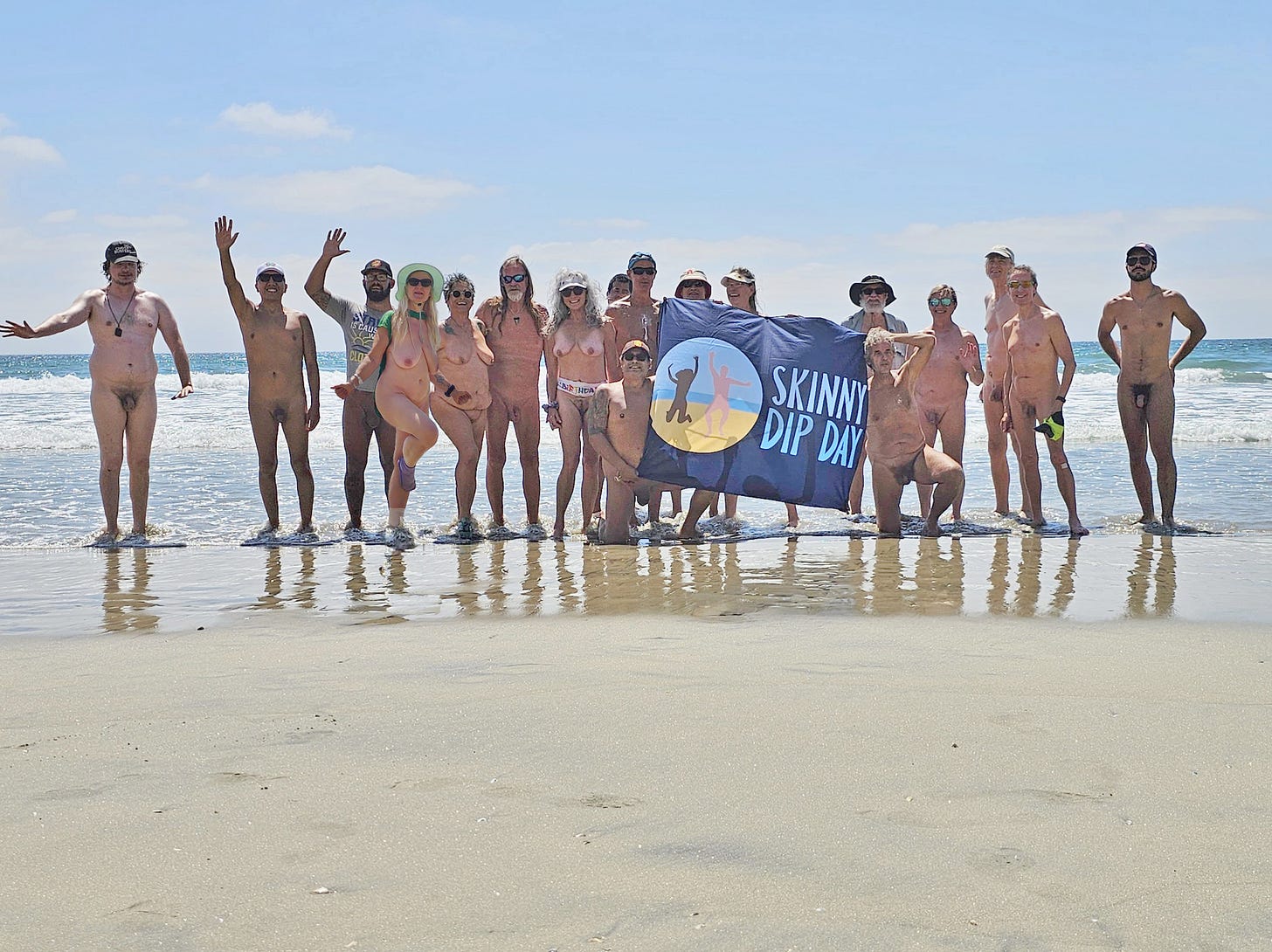 A group of nude beachgoers stand in the shallow surf at Black’s Beach in San Diego, smiling and posing with a Skinny Dip Day flag. The group includes members of San Diego Camping Bares, Naturists in the OC, and other local skinny dippers. A group of nude beachgoers stand in the shallow surf at Black’s Beach in San Diego, smiling and posing with a Skinny Dip Day flag. The group includes members of San Diego Camping Bares, Naturists in the OC, and other local skinny dippers.