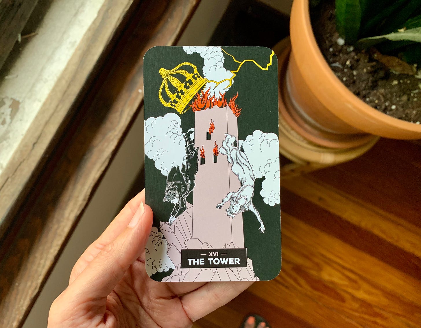 A hand is holding a tarot card next to a window with a wooden sill, and a plant in a large terra cotta pot. In the image, a tower is being struck by lightening and on fire at the top. flames are coming out the windows and two people are flying with calm facial expressions out of it. There is a crown on the tower that is being destabilized in the ruckus. The image is drawn by Xaviera Lopez. A hand is holding a tarot card next to a window with a wooden sill, and a plant in a large terra cotta pot. In the image, a tower is being struck by lightening and on fire at the top. flames are coming out the windows and two people are flying with calm facial expressions out of it. There is a crown on the tower that is being destabilized in the ruckus. The image is drawn by Xaviera Lopez.