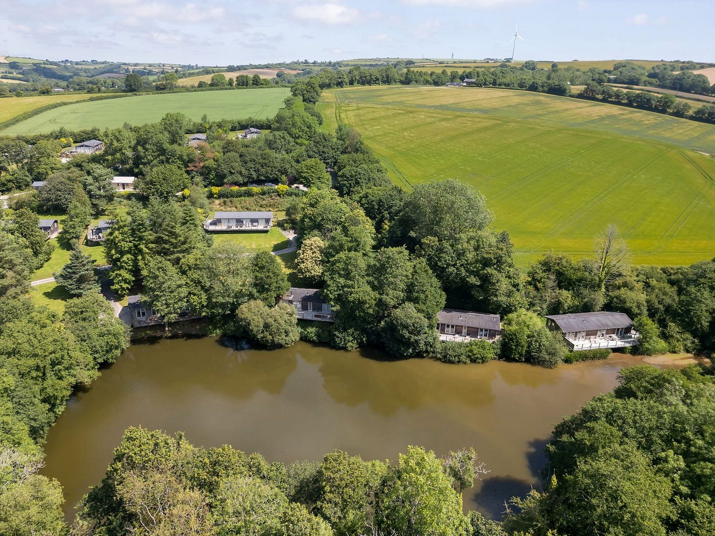 A lake surrounded by trees and houses with fields and hills in the background at 4 Water's Edge in Lanreath