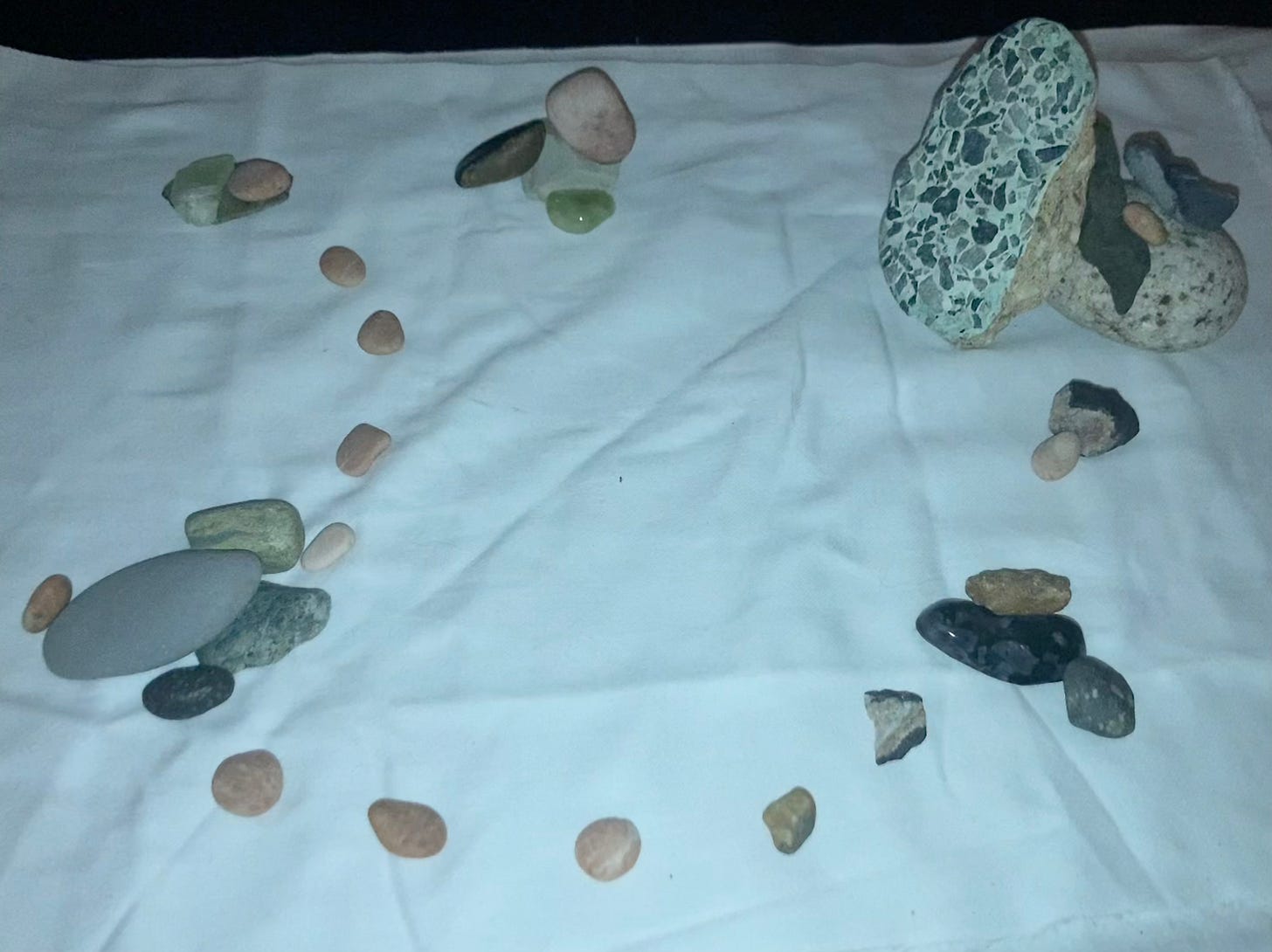 A photo of a scene of rocks, a small pink rock wanders and climbs through many clusters of rocks. Or, it could be read as a a path of rocks following one another and traveling across clusters of rocks. The rocks are pictured on a wrinkled piece of white fabric.