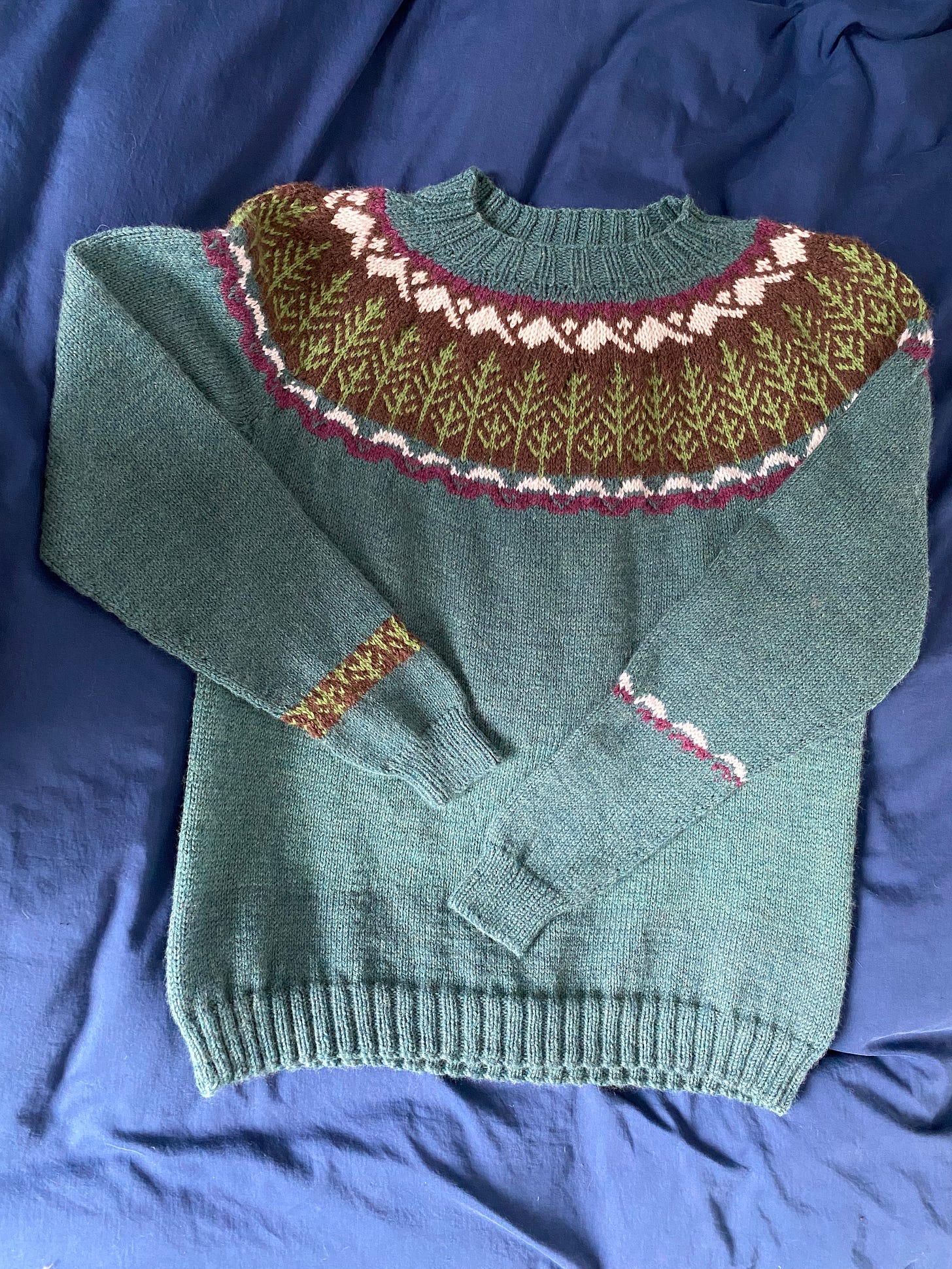 A teal sweater displayed on a navy blue background. The upper yoke has a multicolored pattern of green trees and brown snow-capped mountains.