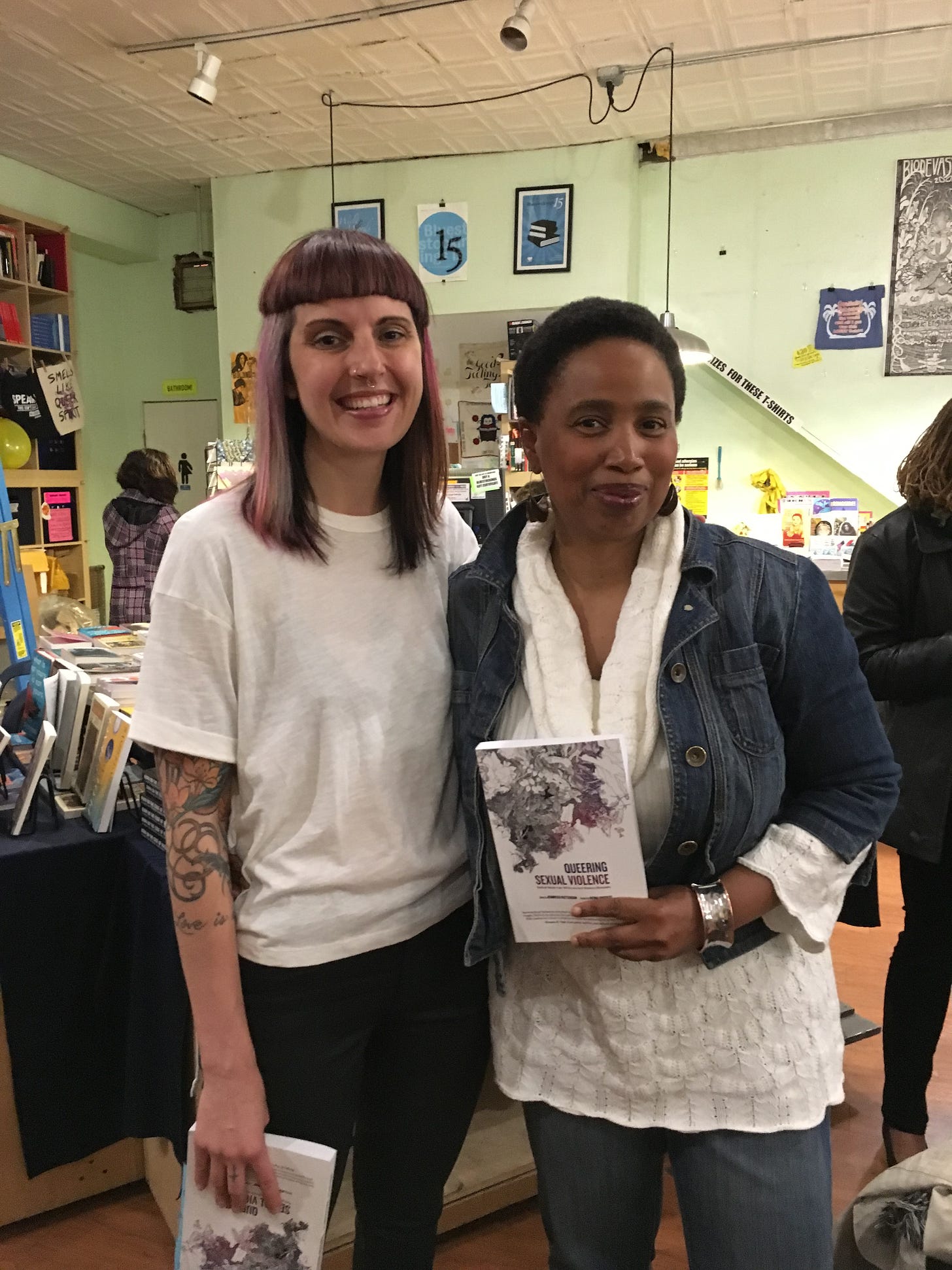 Left to right: Jennifer Patterson and Aishah Shahidah Simmons standing together smiling at Bluestockings Bookstore in May 2016; Aishah holds a copy of Queering Sexual Violence.