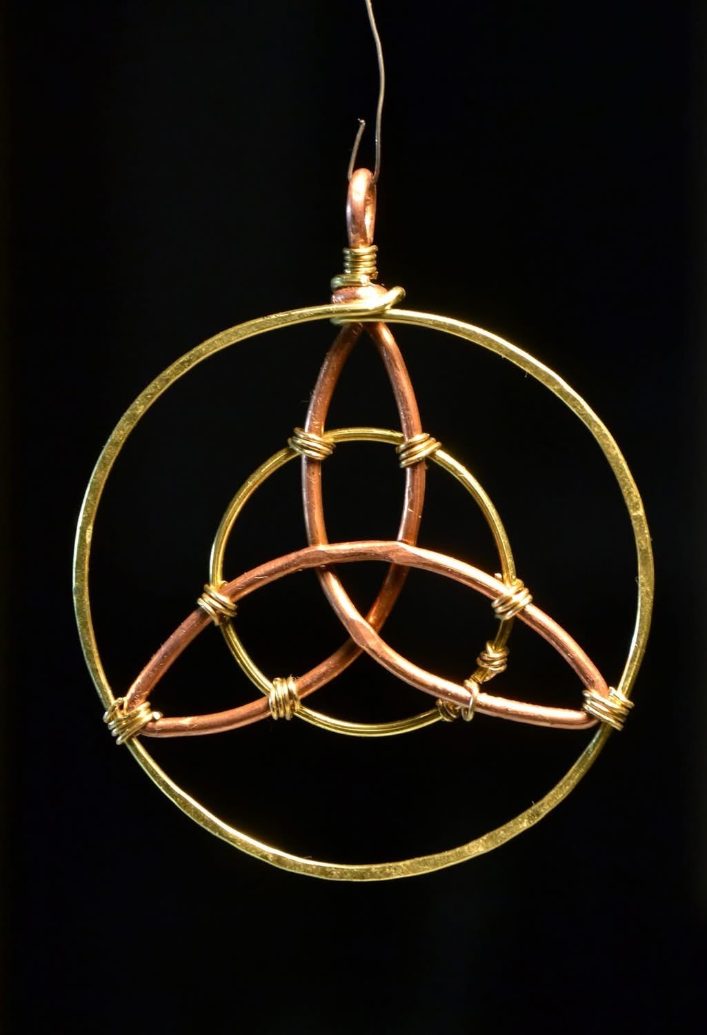 Close-up of a handmade Triquetra pendant crafted from twisted copper and brass wire, formed into an interwoven three-pointed knot inside a circular frame, suspended against a black background.