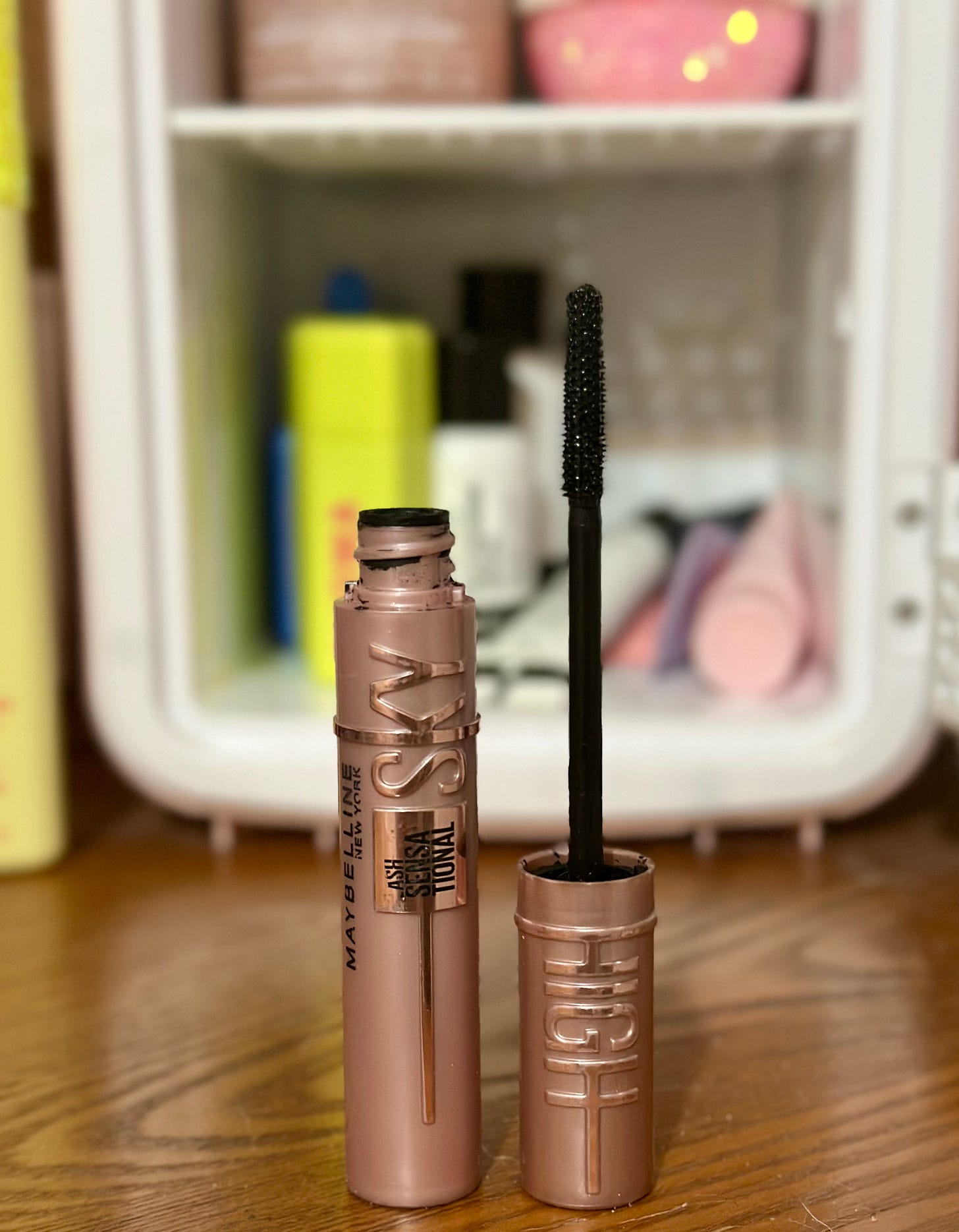 Maybelline Sky High mascara in metallic rose gold packaging. The open tube stands next to the brush wand, which shows the spiky, thin, black plastic bristles. In the blurry background are assorted beauty products including a small pink fridge.